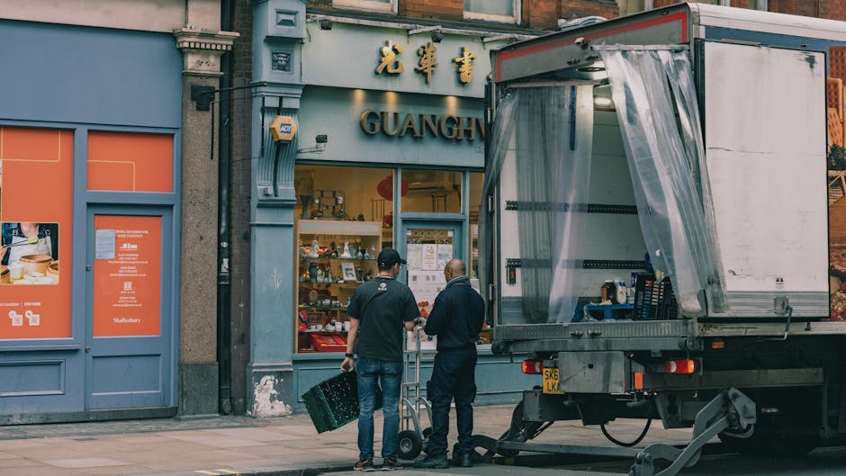 Two removal workers from Man and Van West Kensington are preparing for a home relocation outside a building with the sign 'GUANG,' which appears to be a shop or restaurant. One worker, wearing dark clothing and a cap, holds a crate, while the other, dressed in black, is standing nearby with a hand on a trolley. A large, open-sided delivery truck with a transparent plastic curtain on the side is parked on the pavement directly in front of the premises, indicating the loading process for furniture, boxes, or appliances. Inside the truck's cargo area, visible packing materials such as bubble wrap and cardboard boxes suggest preparation for transport. The surrounding environment includes a building façade with decorative architecture and signage in Chinese characters, reflecting an urban setting suitable for house and business moving logistics. This scene illustrates the initial stages of packing and loading during a professional removal, consistent with services offered by Man and Van West Kensington.