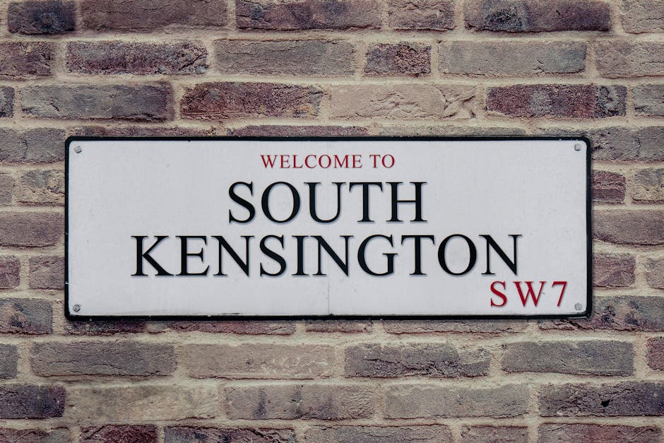 A multi-storey red brick building with white window frames and decorative white vertical accents on the corner, situated on a residential street with other similar buildings. The street features a bla