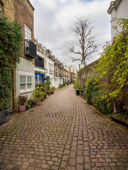 A narrow cobbled street in West Kensington lined with residential buildings, including a mix of brick and painted facades with small balconies and windows. Potted plants and shrubs are placed along the base of the buildings on the left side, while a tall, leafless tree stands at the end of the street against a cloudy sky. On the right side, a high stone wall with green foliage partially covers its surface. The image captures a quiet, urban residential area suitable for home relocations and furniture transport, with no visible moving equipment or personnel present, reflecting the typical environment for house removal services like those offered by Man and Van West Kensington.