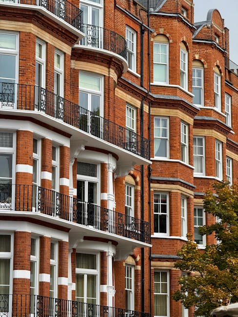 A close-up view of a red brick residential apartment building with white masonry accents and black wrought iron balcony railings running along multiple floors. The building features large sash windows with white frames, some open to reveal curtains inside. On the right side, a section of the building extends higher and has rounded architectural elements. In the foreground, part of a tree with green and yellow leaves is visible, indicating a mild weather day. The overall scene is well-lit, capturing the exterior facade typical of West Kensington’s urban housing, relevant to services provided by Man and Van West Kensington for home relocation and furniture transport during moving or packing and moving operations.