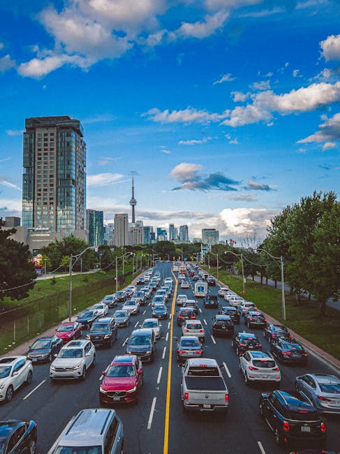 A multi-lane urban street scene during daylight hours shows a variety of vehicles including cars, vans, and trucks moving in both directions. The street is lined with green trees and street lamps along the pavement. In the background, there are modern high-rise buildings and a distinctive radio tower set against a partly cloudy blue sky. The scene captures typical city traffic, possibly during a busy period, with some vehicles appearing to be in the process of moving or stopping. The image reflects a city environment suitable for house removals and logistics planning, as seen in services like Man and Van West Kensington, supporting efficient furniture transport and home relocation processes.