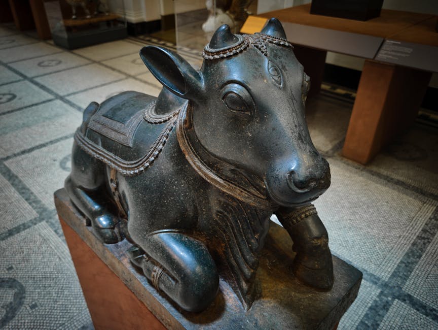 A detailed photograph of a dark metal sculpture depicting a stylized animal figure resembling a cow or buffalo, seated on a rectangular wooden pedestal. The sculpture features intricate decorative elements, including a headdress or jewelry style adornments around the head and neck area, with patterns resembling beads or chains. The animal's eyes are large and rounded, with a gentle facial expression. Its body shows carved details representing folds and textures, and the front legs are positioned close together as it sits on the pedestal. The sculpture is placed on a patterned beige carpet, with a wooden table visible in the background, indicating an indoor setting that could be a museum or a private collection. The lighting is soft, highlighting the metallic sheen and the crafted details of the piece.