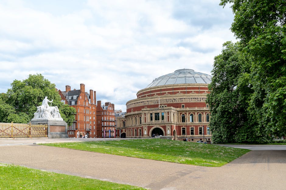 Photograph of the Royal Albert Hall in London, taken from a nearby park. The historic building features a large, rounded brick and stone facade with arched windows and an iconic circular roof. In the foreground, there is a paved walkway and well-maintained grass with scattered white flowers. To the left, a white marble statue depicting classical figures sits atop a pedestal, partially obscured by green trees. Large leafy trees surround the scene, creating a natural frame around the historic venue. The sky is overcast with light grey clouds, providing even, natural lighting over the area. The scene captures the exterior surroundings during daytime, with no visible signage or vehicles directly in front of the building. This setting aligns with a location where house removals or furniture transport to a cultural site could occur, and the image supports content related to moving or relocation services focused on central London.