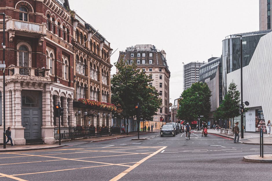A daytime street scene in West Kensington showing a wide road with marked pedestrian crossings and several trees lining the pavement. On the left, there is a historic red-brick building with ornate white stone detailing, balconies, and flower boxes on the windowsills, indicating a residential or commercial property. To the right, modern office buildings with sleek glass facades are visible. There are a few pedestrians walking along the sidewalk, some waiting at traffic lights, and a vehicle moving through the intersection. In the foreground, a man and woman are seen near the curb, possibly preparing for a house or office move, with one person carrying a box wrapped in plastic or fabric and a trolley loaded with packing materials nearby, reflecting the process of packing and loading items for relocation. The scene conveys an active urban environment typical of a well-developed area in West Kensington, suitable for professional removals and furniture transport services like those provided by Man and Van West Kensington.