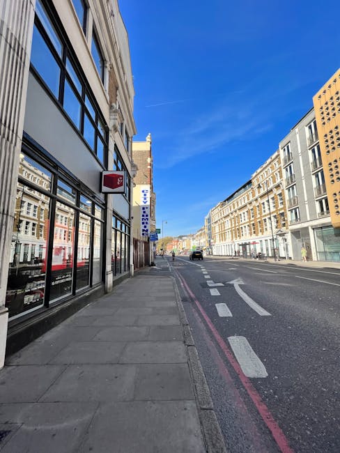 An urban street scene showing the exterior of a building with large glass windows on the left side, displaying shelves and items inside. Outside, the sidewalk extends along the building, with a few boxes and packing materials placed temporarily near the storefront. The street features a red line along the curb indicating parking restrictions, with a few vehicles driving or parked further down the road. On the right side, modern multi-storey buildings with a mix of light and dark facades line the street. The clear blue sky overhead suggests good weather. This scene represents a typical location for a house or office relocation, where packing materials and furniture might be organized on the pavement before loading into a van. Man and Van West Kensington often assist with such moving processes, ensuring smooth furniture transport and packing for clients in the North End Road area.