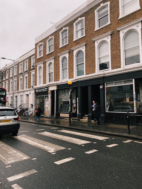 A daytime street scene in West Kensington showing a wide road with marked pedestrian crossings and several trees lining the pavement. On the left, there is a historic red-brick building with ornate white stone detailing, balconies, and flower boxes on the windowsills, indicating a residential or commercial property. To the right, modern office buildings with sleek glass facades are visible. There are a few pedestrians walking along the sidewalk, some waiting at traffic lights, and a vehicle moving through the intersection. In the foreground, a man and woman are seen near the curb, possibly preparing for a house or office move, with one person carrying a box wrapped in plastic or fabric and a trolley loaded with packing materials nearby, reflecting the process of packing and loading items for relocation. The scene conveys an active urban environment typical of a well-developed area in West Kensington, suitable for professional removals and furniture transport services like those provided by Man and Van West Kensington.
