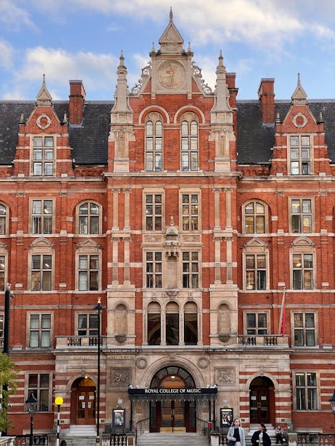 A historic red-brick building with intricate stone detailing and multiple large windows, situated on a city street under a blue sky. The building features ornate gables, decorative spires, and a centr
