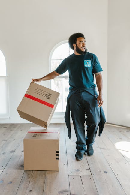 A man with a dark curly beard and short curly hair, wearing a navy blue t-shirt with a light blue logo on the chest and dark trousers, is inside a bright, modern room with white walls and large arched windows. He is holding a medium-sized cardboard box wrapped with a red tape strip in his right hand, while standing next to two other cardboard boxes placed on a light wooden floor. The boxes appear prepared for a home relocation or furniture transport, with the room indicating the early stages of packing and moving. The man is positioned near a doorway leading to another room, and natural light from the windows illuminates the scene, highlighting the interior space. Man and Van West Kensington, a professional removals service, is involved in the logistics of packing and loading materials, contributing to a smooth furniture transport and house move process.