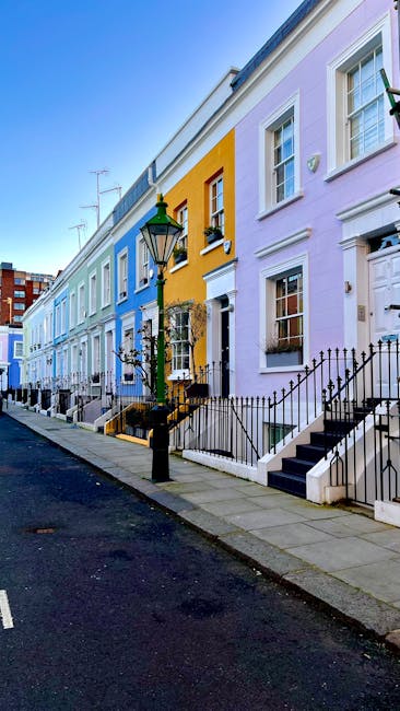 A row of colorful terraced houses on a residential street in West Kensington, with pastel facades in shades of light blue, yellow, pink, and lavender, each with a small front garden enclosed by black wrought iron railings, stone steps leading up to white front doors, and white-framed windows. A classic black lamppost stands on the pavement near the houses, with a green decorative lantern on top. The street is empty, with a dark asphalt road and a concrete sidewalk, under a clear blue sky. This scene exemplifies the residential architecture typical of West Kensington, often associated with house removals and home relocation services. The image provides a visual context for the logistical process of furniture transport and moving logistics, highlighting the urban environment where professional removals companies like Man and Van West Kensington may operate, especially for same-day removals and packing and moving services in the area.