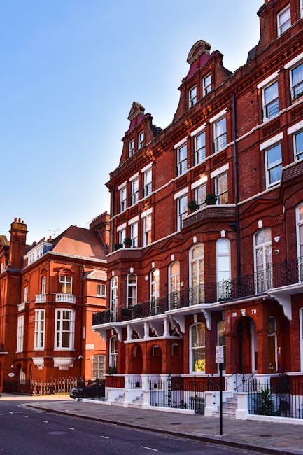 A row of colorful terraced houses on a residential street in West Kensington, with pastel facades in shades of light blue, yellow, pink, and lavender, each with a small front garden enclosed by black wrought iron railings, stone steps leading up to white front doors, and white-framed windows. A classic black lamppost stands on the pavement near the houses, with a green decorative lantern on top. The street is empty, with a dark asphalt road and a concrete sidewalk, under a clear blue sky. This scene exemplifies the residential architecture typical of West Kensington, often associated with house removals and home relocation services. The image provides a visual context for the logistical process of furniture transport and moving logistics, highlighting the urban environment where professional removals companies like Man and Van West Kensington may operate, especially for same-day removals and packing and moving services in the area.